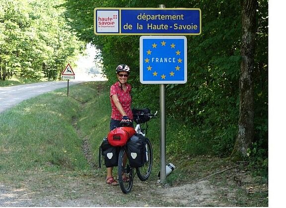 Fahrradfahrerin steht mit bepacktem Fahrrad unter "Frankreichschild"