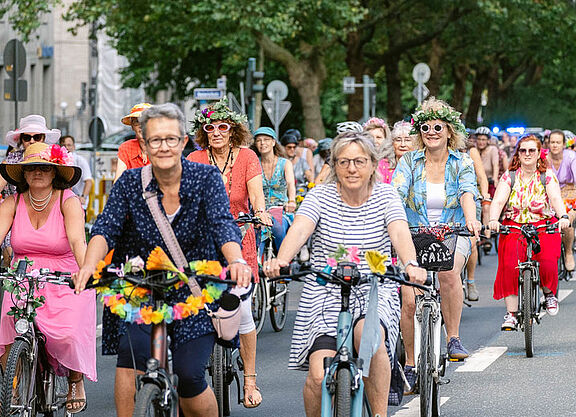 Bunt gekleidete Frauen auf geschmückten Fahrrädern lachend auf einer Straße radelndf ei