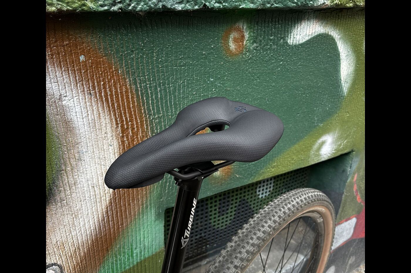 Schwarzer Fahrradsattel mit mittlerer Aussparung, montiert an einem Fahrrad vor einer bemalten Wand.