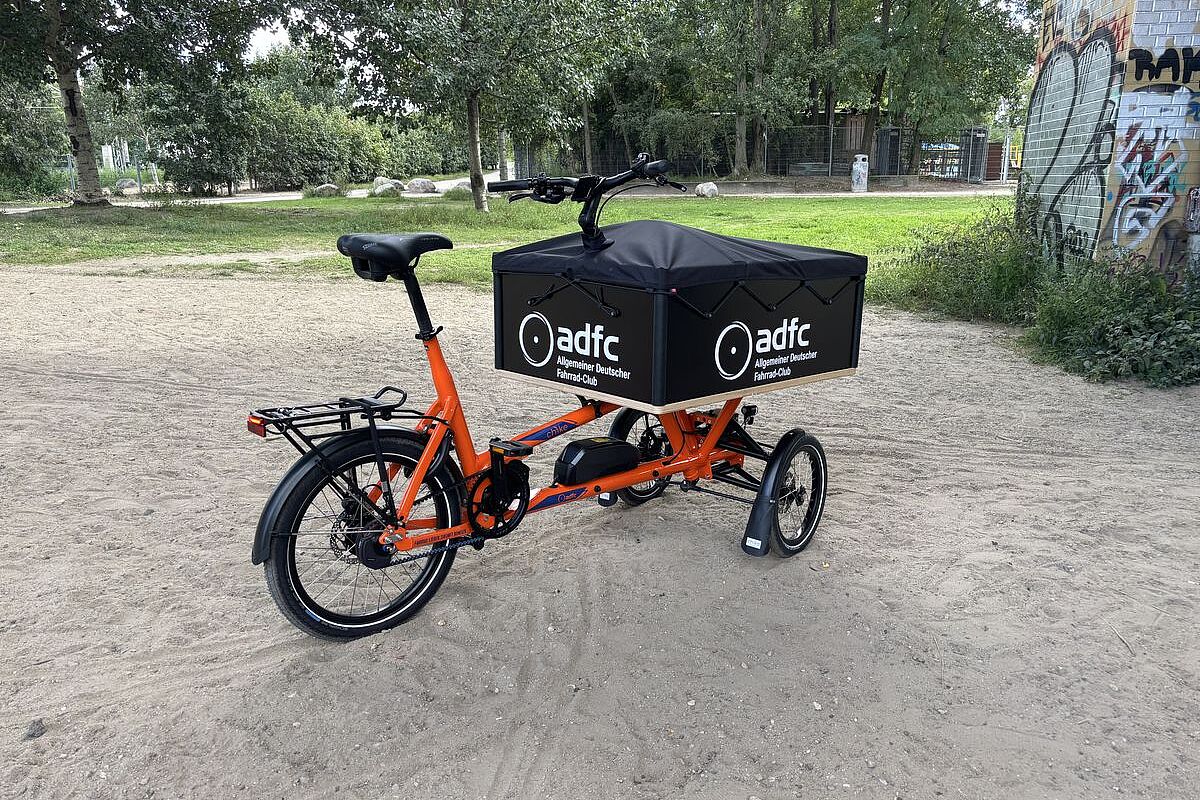 Orangefarbenes dreirädriges Lastenrad mit schwarzer ADFC-Transportbox steht auf einem sandigen Weg im Grünen.